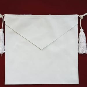 May include: A white apron with a triangular flap at the top. White tassels hang from each side, attached by white cords. The apron is set against a dark red background.