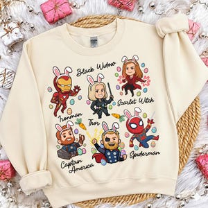 May include: Cream-colored sweatshirt featuring cartoon illustrations of Marvel characters Iron Man, Black Widow, Scarlet Witch, Thor, Captain America, and Spiderman, each wearing bunny ears. The names of the characters are written above or below them.