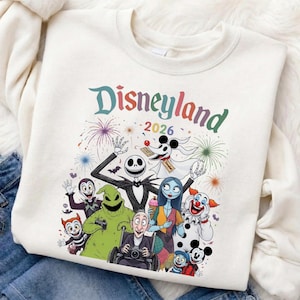 May include: Cream-colored sweatshirt featuring a colorful graphic of Disneyland characters, including Jack Skellington, Sally, and Zero. The word "Disneyland" is written in rainbow colors above the characters, with "2026" below. Fireworks are in the background.