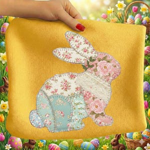 Puede incluir: Un cojín amarillo con un diseño de conejo de patchwork. El conejo está hecho de tela floral en rosa, blanco y azul, con un detalle de encaje. El cojín está sostenido por una mano con esmalte de uñas rojo. Tema de Pascua.