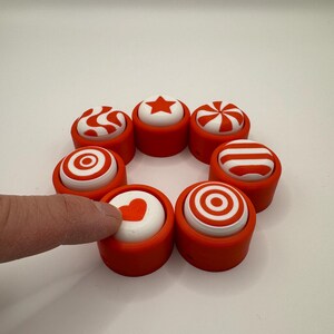 May include: Seven red and white plastic buttons with different designs, including a heart, a star, a swirl, stripes, and circles. The buttons are arranged in a circle on a white surface.
