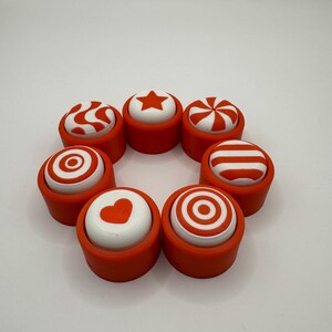 May include: Eight orange and white push buttons with different designs, including a star, heart, stripes, and swirls. The buttons are arranged in a circle.