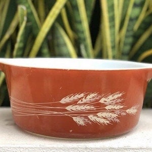 May include: A vintage, rust-colored Pyrex bowl with a white interior. The exterior features a wheat pattern in white. The bowl has a rounded shape with a flared rim and two small handles. A classic piece for kitchenware.