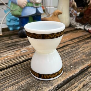 May include: A white, hourglass-shaped egg cup with black and gold decorative bands. The vintage kitchenware item is made of opaque glass and sits on a weathered wooden surface. The egg cup has a wide top and base, with a narrower middle section.