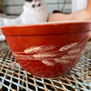 May include: A red mixing bowl with a white wheat design. The bowl is sitting on a metal surface.