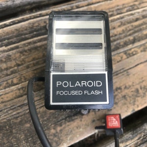 May include: A black Polaroid Focused Flash unit with a silver reflector and a black cord. The unit has the word "POLAROID" and "FOCUSED FLASH" printed on a black label. A red plug is attached to the cord.