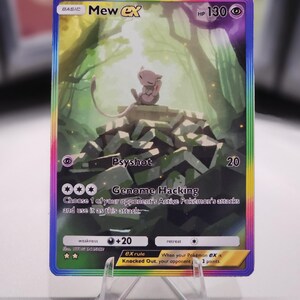 May include: A Pokemon trading card featuring Mew EX, a pink and white mythical Pokemon. The card has a rainbow border and a green and white background. The card's text reads "BASIC Mew EX HP 130 Psyshot 20 *** Genome Hacking Choose 1 of your opponent's Active Pokemon's attacks and use it as this attack weakness +20 retreat ex rule When your Pokemon ex is Knocked Out, your opponent takes 2 prize cards."