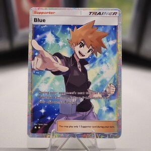 May include: A Pokemon trading card featuring Blue, a character from the Pokemon franchise. The card is a supporter card and has a blue, pink, and white background with stars. The text on the card reads "Supporter, Blue, Trainer, During your opponent's next turn, all of your Pokemon take -10 damage from attacks from your opponent's Pokemon. You may play only 1 Supporter card during your turn."