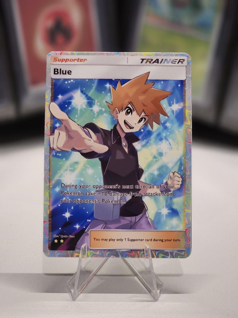 May include: A Pokemon trading card featuring Blue, a character from the Pokemon franchise. The card is a supporter card and has a blue, pink, and white background with stars. The text on the card reads "Supporter, Blue, Trainer, During your opponent's next turn, all of your Pokemon take -10 damage from attacks from your opponent's Pokemon. You may play only 1 Supporter card during your turn."