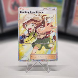 May include: A Pokemon trading card featuring a young girl in a green shirt and brown hat holding a camera. The card is titled "Budding Expeditioner" and is a Trainer card. The text on the card reads "Put your Mew ex in the Active Spot, into your hand." and "You may play only 1 Supporter card during your turn."