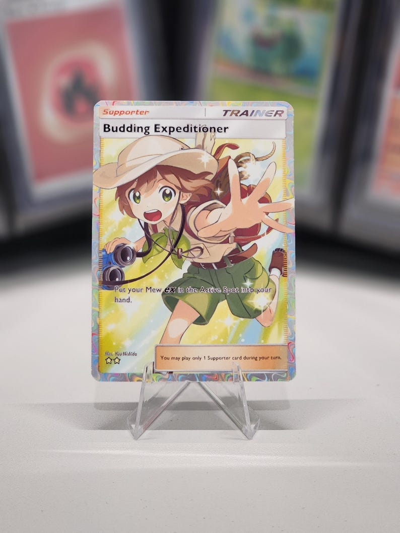 May include: A Pokemon trading card featuring a young girl in a green shirt and brown hat holding a camera. The card is titled "Budding Expeditioner" and is a Trainer card. The text on the card reads "Put your Mew ex in the Active Spot, into your hand." and "You may play only 1 Supporter card during your turn."