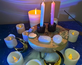 Può includere: Una scena mistica con candele accese, tra cui due alte in supporti neri e diverse candele più piccole a batteria. Una piattaforma di legno contiene cristalli e altri oggetti decorativi. Accenti dorati a forma di luna crescente e uno sfondo blu stellato completano l'esposizione.
