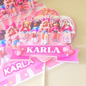 May include: A pink and white cake topper with the name "KARLA" in large, bold letters. The topper features several doll figures wearing pink tops and denim shorts. The overall design is bright and cheerful, perfect for a birthday celebration.
