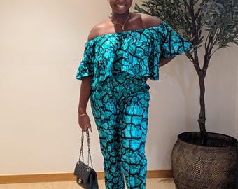 May include: A woman wearing a turquoise off-the-shoulder jumpsuit with a black and white pattern. She is wearing black sandals and carrying a black handbag.