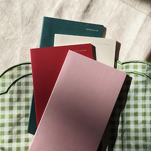 May include: A stack of four rectangular grid notebooks in shades of dark green, cream, deep red, and light pink. Each notebook cover features the text Grid Notebook embossed in the upper right corner, arranged in a staggered, overlapping vertical pile.