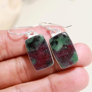 May include: A pair of dangle earrings featuring rectangular, polished ruby zoisite stones with mottled green and deep magenta patterns. Each stone is set in a simple silver-toned metal frame and attached to a curved silver-toned ear wire hook.