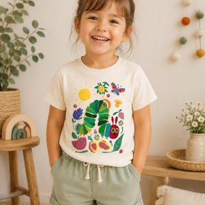May include: A young child wearing a cream-colored t-shirt with a colorful illustration of a caterpillar, sun, flowers, and butterfly. The child is also wearing green shorts. The image is taken indoors.