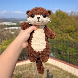 May include: A handmade crochet otter plushie in shades of brown and cream. The otter has a detailed face with black eyes, a cream-colored muzzle, and white whiskers. The plushie is held up against a blurred outdoor background.