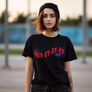 May include: A woman wearing a black t-shirt with the text "HEY-EY-EY" in red and a blue buffalo silhouette. She is also wearing a black beanie and black jeans.