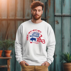 May include: White sweatshirt with a blue and red graphic of a buffalo inside a football helmet. The graphic says "Hey-ey-ey-ey Let's Go Buffalo".