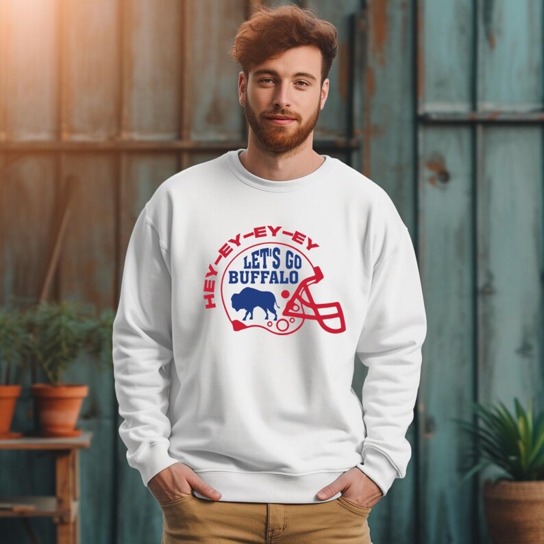 May include: White sweatshirt with a blue and red graphic of a buffalo inside a football helmet. The graphic says "Hey-ey-ey-ey Let's Go Buffalo".