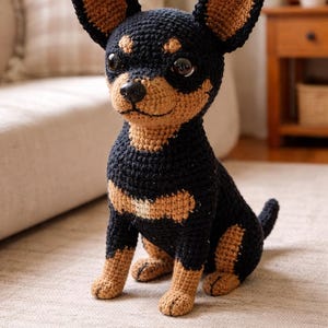 May include: A crocheted dog figurine in a sitting position. The dog is primarily black with tan markings on its ears, paws, and chest. It has large, dark eyes and a small, black nose. The dog is sitting on a light-colored rug.