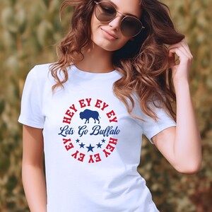 May include: White t-shirt with a red, white, and blue circle graphic. The graphic features a blue buffalo silhouette, the text "Let's Go Buffalo", and five white stars. The text "HEY EY EY EY HEY" is written in a circle around the buffalo and stars.