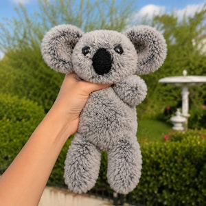 May include: A plush koala toy crafted from soft, textured grey yarn. The stuffed animal features large, rounded ears, a prominent black nose, and dark, round eyes. The material has a fuzzy, shaggy appearance throughout the entire body.