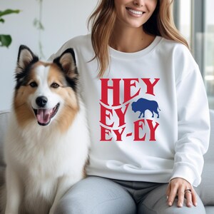 May include: White sweatshirt with red text that reads "HEY EY EY-EY" and a blue buffalo silhouette.