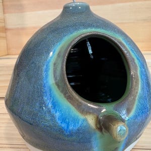 May include: A spherical ceramic birdhouse with a glossy glaze in shades of deep blue, teal, and green. It features a large circular opening on the front, a small perch extending below the opening, and a metal hanging wire at the top.