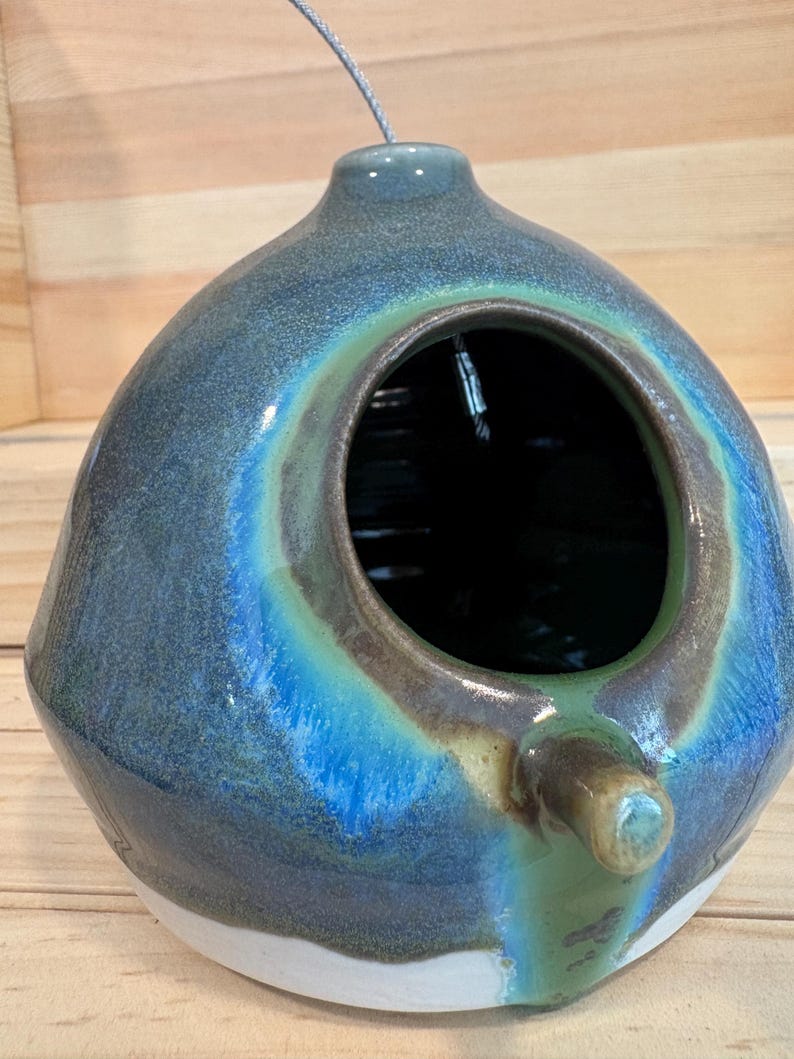 May include: A spherical ceramic birdhouse with a glossy glaze in shades of deep blue, teal, and green. It features a large circular opening on the front, a small perch extending below the opening, and a metal hanging wire at the top.