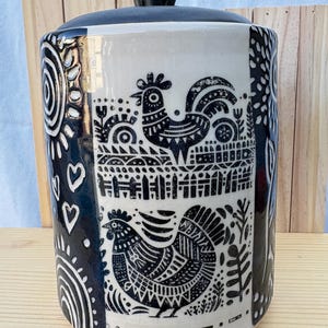 May include: A cylindrical ceramic canister with a black lid, featuring a white surface decorated with black ink illustrations of two roosters, a picket fence, hearts, and sunburst patterns. The design is detailed with geometric lines and floral motifs.