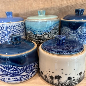 May include: Five ceramic lidded jars with various blue and white patterns. The collection includes designs featuring circles, swirling waves, and floral motifs. Each jar has a matching lid with a small knob handle, showcasing a mix of solid blue, speckled, and intricate hand-drawn details on a cylindrical body.
