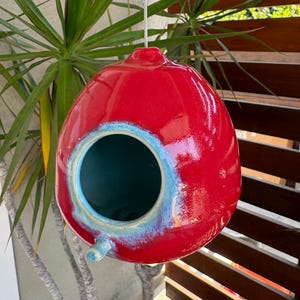 May include: A glossy, spherical ceramic birdhouse featuring a vibrant red exterior finish. The circular entrance is framed by a light blue, reactive glaze that transitions into the deep red body. A small, cylindrical perch extends below the opening.
