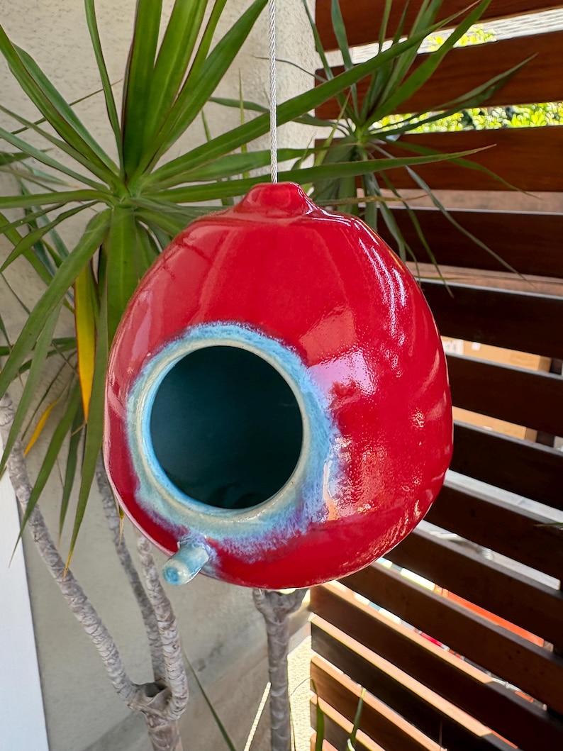 May include: A glossy, spherical ceramic birdhouse featuring a vibrant red exterior finish. The circular entrance is framed by a light blue, reactive glaze that transitions into the deep red body. A small, cylindrical perch extends below the opening.