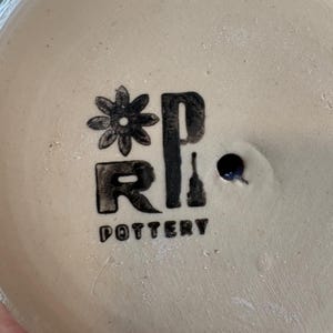 May include: A circular, off-white ceramic base featuring a black stamped logo. The logo includes a stylized flower icon, the letters R and P, and the word POTTERY printed below. A small, dark, circular drainage hole is visible to the right.