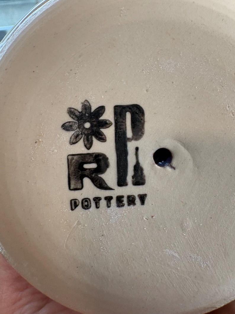 May include: A circular, off-white ceramic base featuring a black stamped logo. The logo includes a stylized flower icon, the letters R and P, and the word POTTERY printed below. A small, dark, circular drainage hole is visible to the right.
