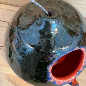 May include: A spherical ceramic birdhouse with a glossy dark green and black crystalline glaze, featuring a bright orange interior and a circular entrance hole. A metal wire extends from the top, and a wooden ruler is visible at the base.