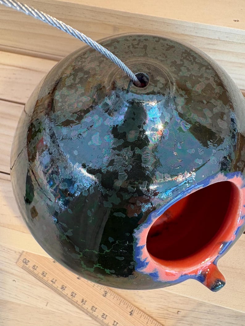 May include: A spherical ceramic birdhouse with a glossy dark green and black crystalline glaze, featuring a bright orange interior and a circular entrance hole. A metal wire extends from the top, and a wooden ruler is visible at the base.