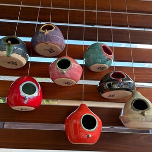 May include: A collection of eight ceramic birdhouses in various colors including red, blue, pink, teal, black, and beige, each featuring a circular opening and a small perch, suspended by thin wires against a horizontal wooden slat background.