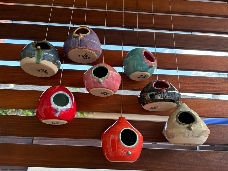 May include: A collection of eight ceramic birdhouses in various colors including red, blue, pink, teal, black, and beige, each featuring a circular opening and a small perch, suspended by thin wires against a horizontal wooden slat background.