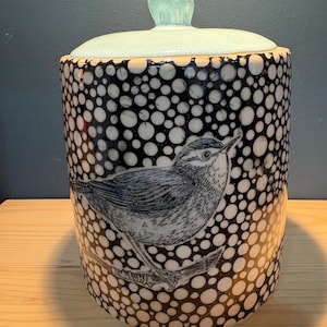 May include: A cylindrical ceramic canister with a lid, featuring a black and white polka dot pattern and a detailed black line drawing of a small bird perched on a branch. The lid is a solid, pale mint green color with a small knob handle.