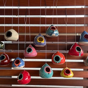 May include: A selection of ceramic birdhouses in a range of colours, such as red, blue, green, and yellow, each with a circular entrance. The birdhouses are hung by strings against a wooden slatted backdrop.