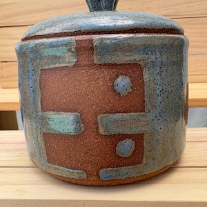 May include: A cylindrical ceramic jar with a matching lid and a knob handle. The exterior features a textured, reddish-brown clay base with a blue, crackled glaze applied in geometric patterns, including two small circular blue accents on the front.