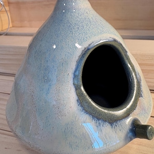 May include: A teardrop-shaped ceramic birdhouse with a glossy, mottled light blue and white glaze. It features a circular entrance hole with a dark, textured rim and a small cylindrical perch extending from the lower right side.