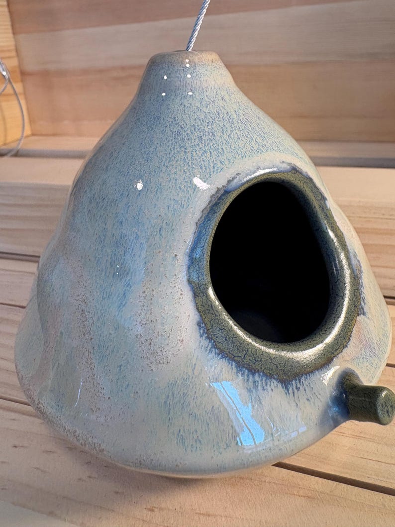 May include: A teardrop-shaped ceramic birdhouse with a glossy, mottled light blue and white glaze. It features a circular entrance hole with a dark, textured rim and a small cylindrical perch extending from the lower right side.