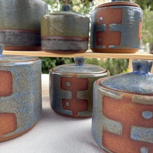 May include: A selection of handmade ceramic jars with lids. The jars are cylindrical, with a blue, brown, and green colour scheme. Some jars have a geometric design in brown. The lids have small knobs. The jars are displayed on a wooden shelf.