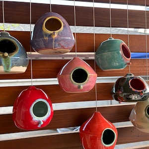 May include: A collection of eight ceramic birdhouses in various colors including purple, red, teal, black, and beige. Each rounded, teardrop-shaped house features a single circular entrance hole and hangs from a thin metal wire.