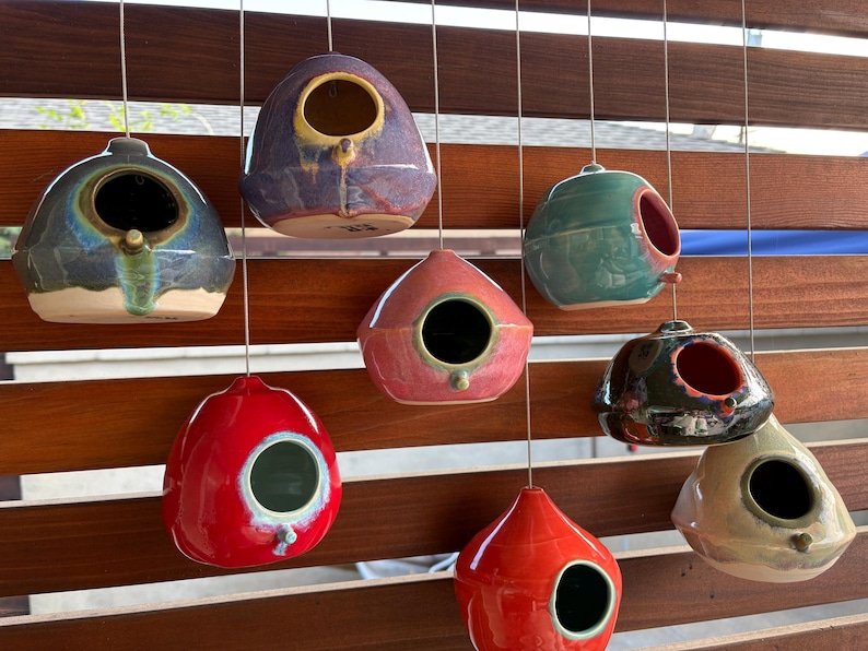 May include: A collection of eight ceramic birdhouses in various colors including purple, red, teal, black, and beige. Each rounded, teardrop-shaped house features a single circular entrance hole and hangs from a thin metal wire.