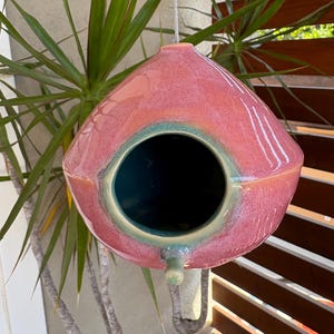 May include: A teardrop-shaped ceramic birdhouse with a glossy pink glaze and a teal-colored interior. The birdhouse features a circular entrance hole, a small protruding perch below the opening, and a metal wire for hanging from the top.